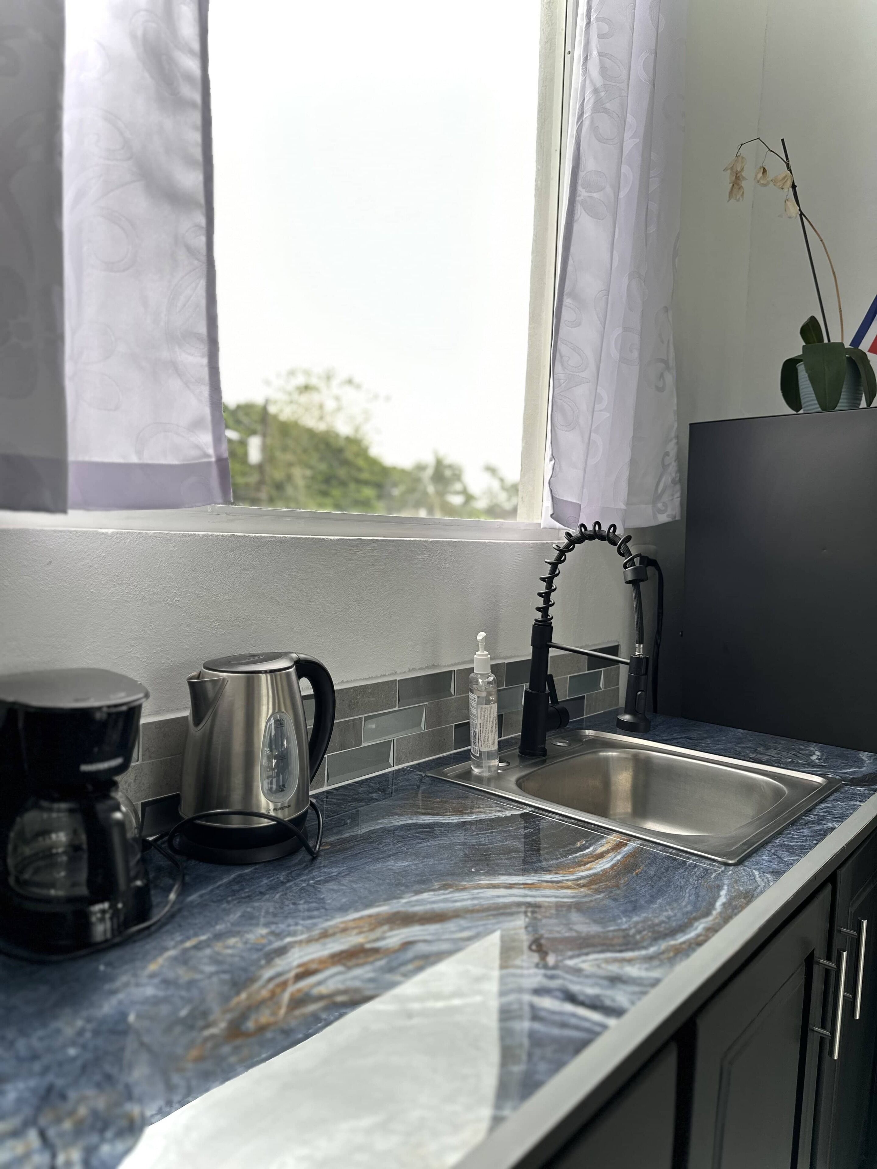 Una cocina con un lavamanos de acero inoxidable, una tetera y una cafetera sobre una encimera de mármol.