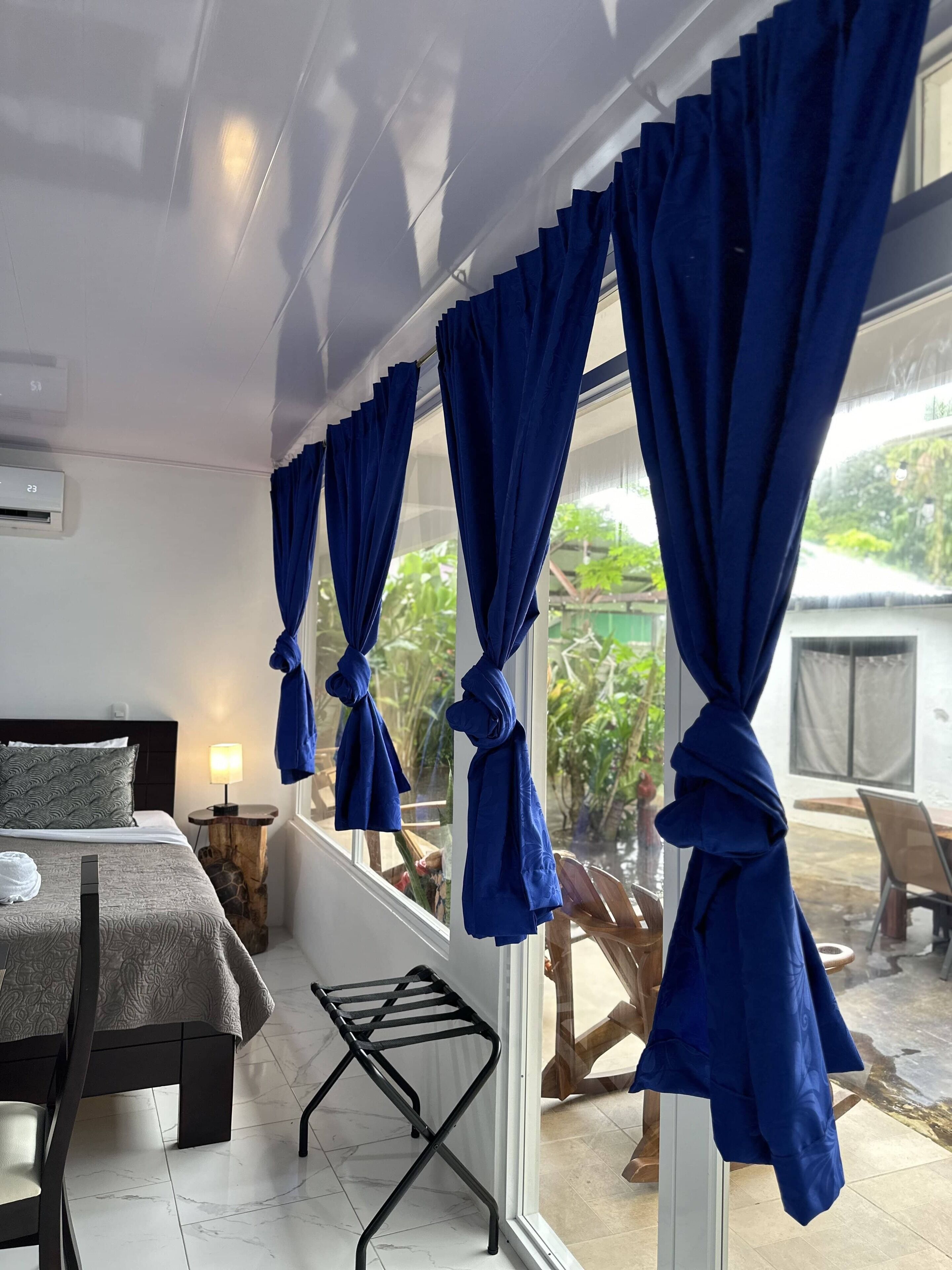Habitación de hotel con una cama, una mesita pequeña, una lámpara y una ventana con cortinas azules recogidas.
