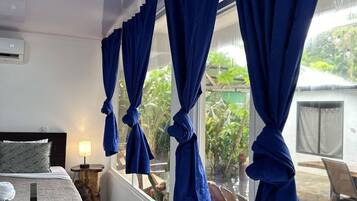 Habitación de hotel con una cama, una mesita pequeña, una lámpara y una ventana con cortinas azules recogidas.