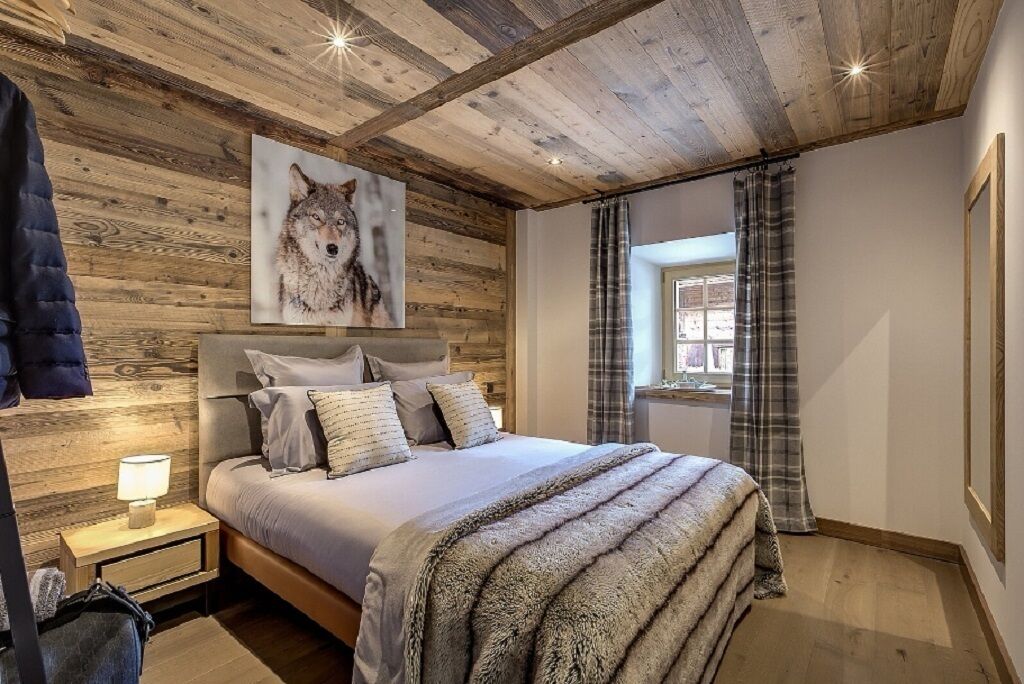 Une chambre avec un plafond en bois, un grand lit recouvert d’une couverture moelleuse, un tableau représentant un loup et une fenêtre avec des rideaux.