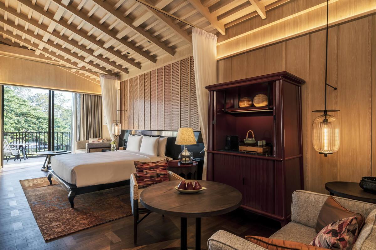 Une chambre d’hôtel moderne dotée d’un grand lit, d’un coin salon avec une table ronde, d’une armoire en bois et d’un balcon offrant une vue sur les arbres.