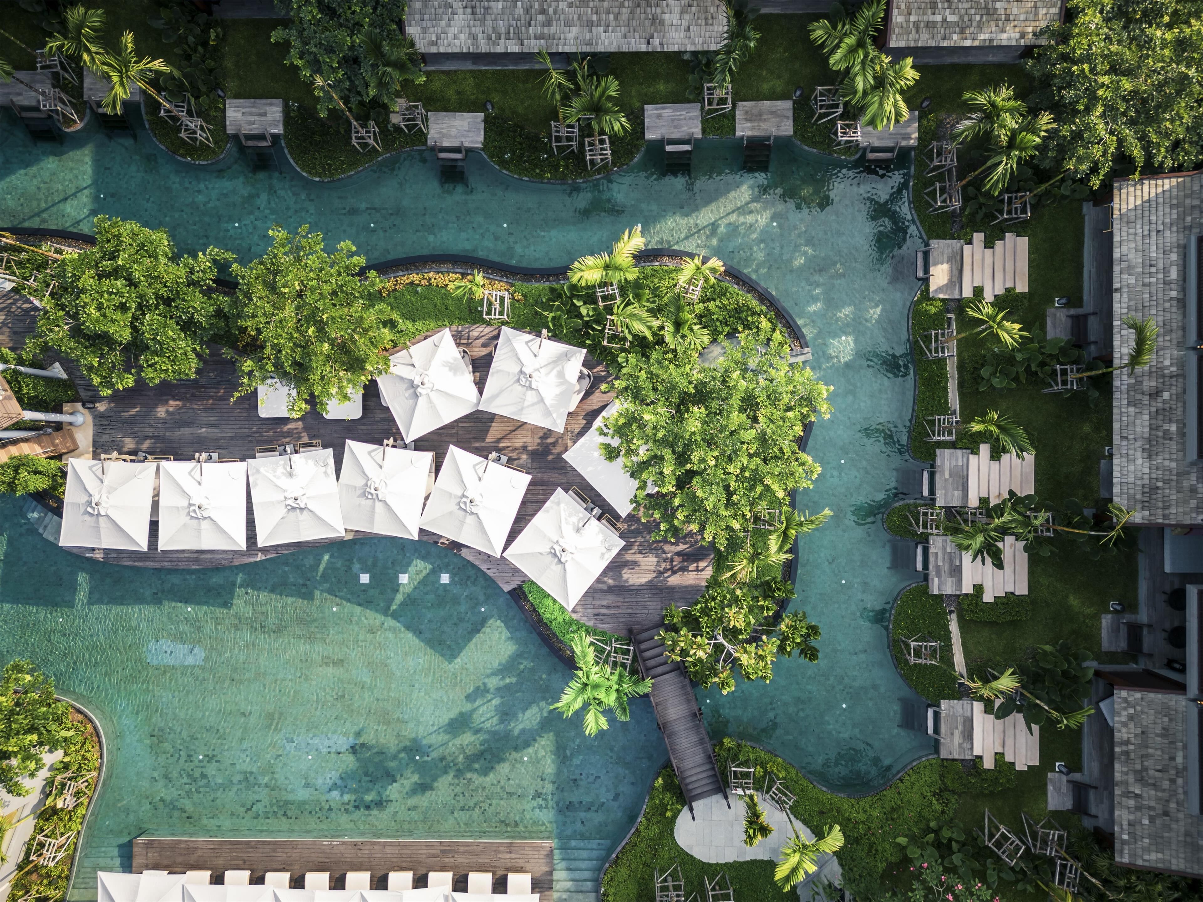 Vue aérienne d’un complexe hôtelier doté d’une piscine centrale, entourée d’espaces de détente avec des parasols et des sièges, ainsi que d’une végétation luxuriante.