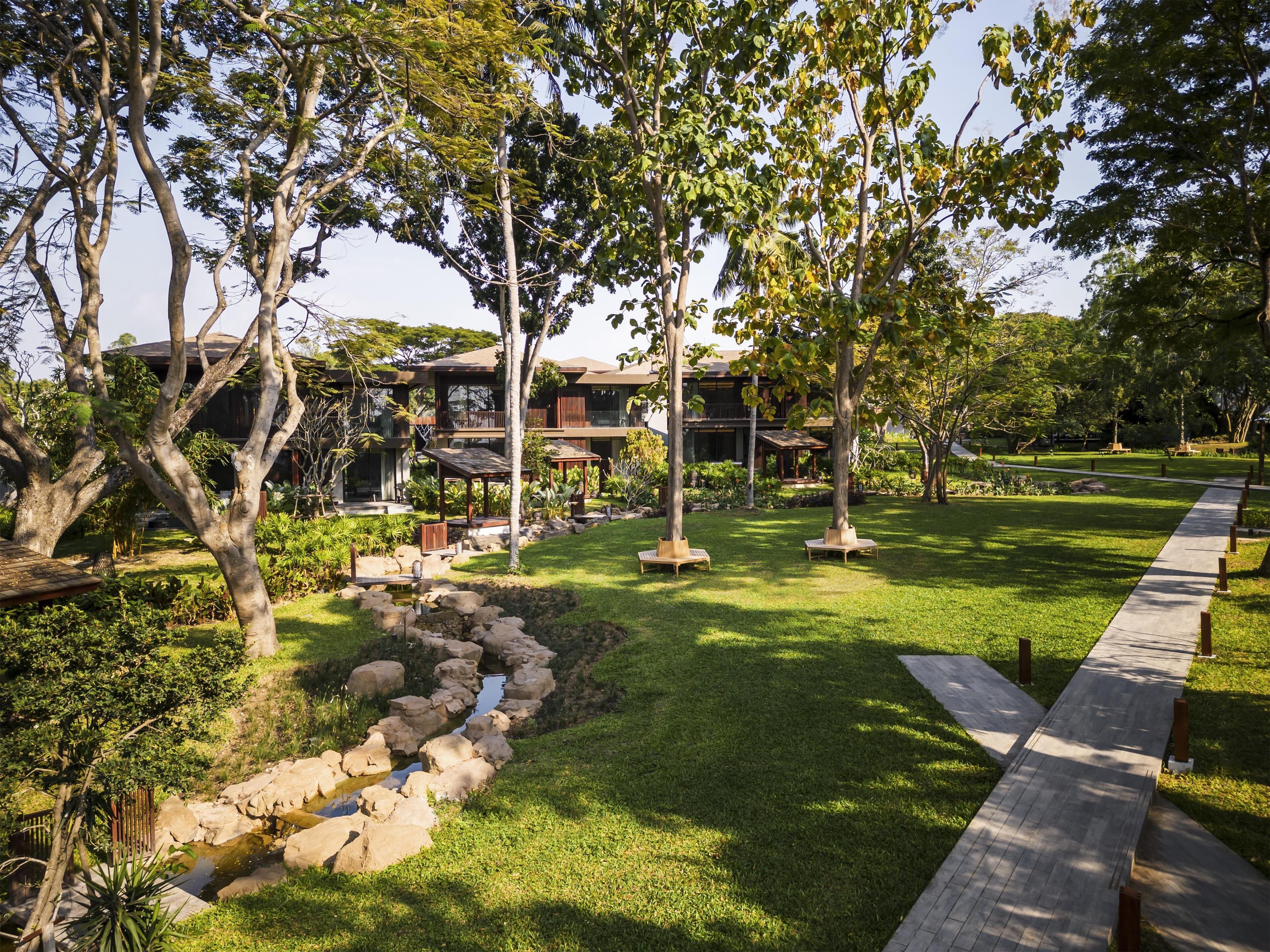 A well-maintained garden with a stone water feature, wooden benches, and a pathway leading to a building with multiple levels and balconies.
