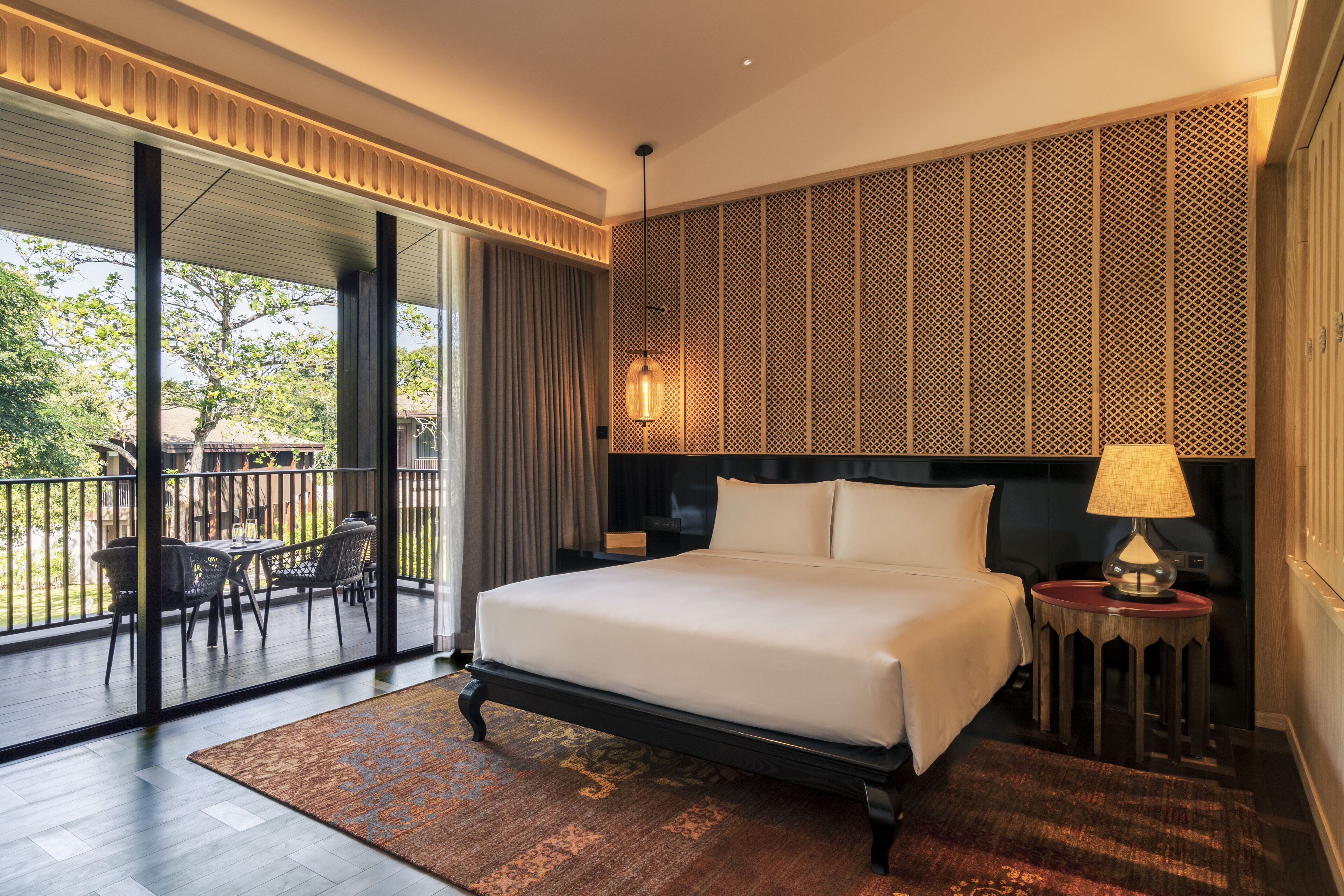 A hotel room with a large bed, a balcony with outdoor furniture, and a patterned wall.