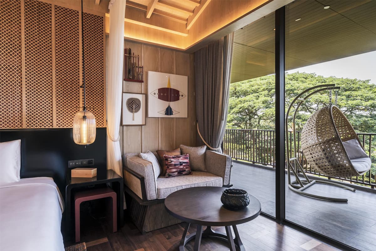 Une chambre d’hôtel moderne, dotée d’un canapé, d’une petite table, d’un fauteuil suspendu et d’une grande fenêtre donnant sur un paysage verdoyant.