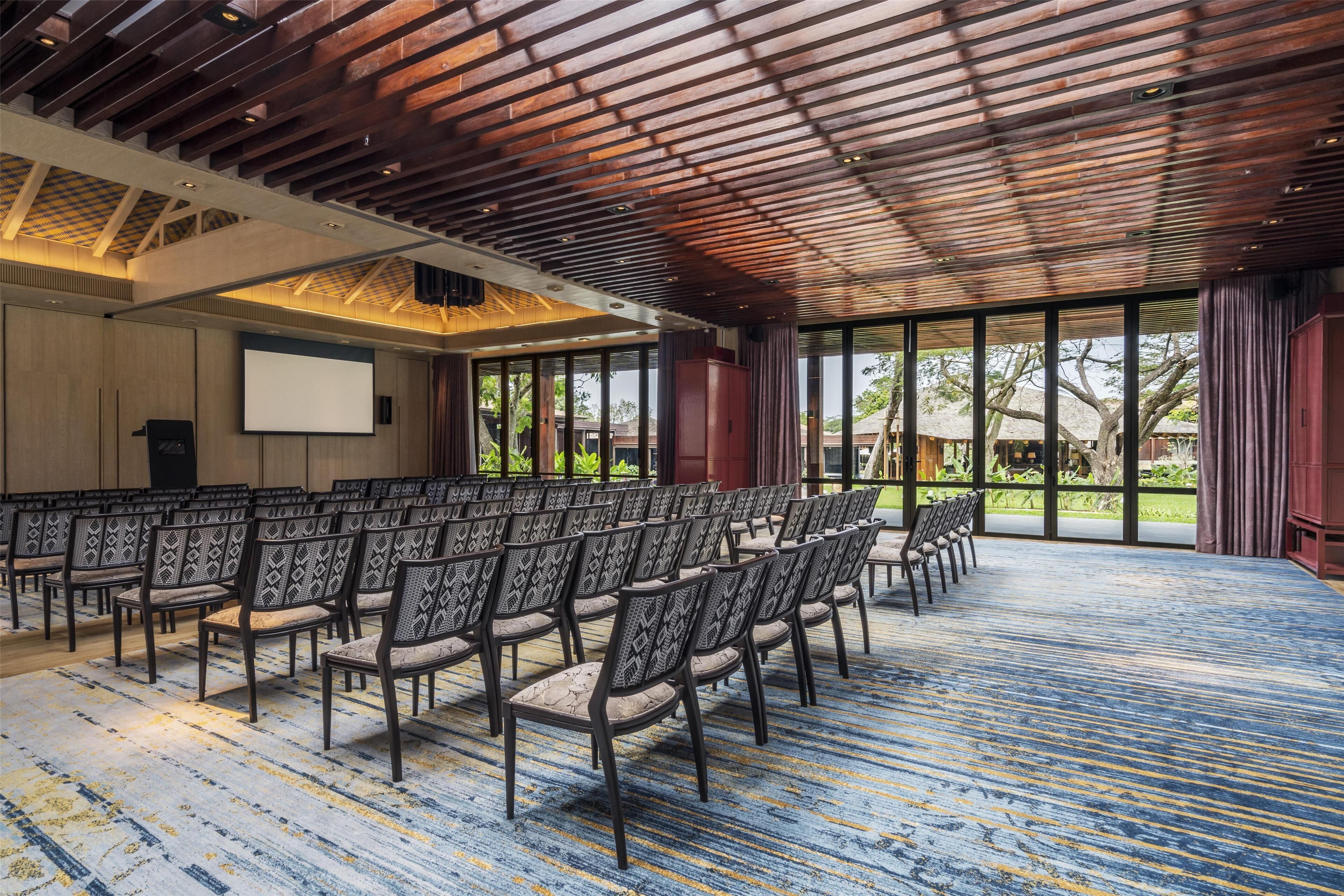 A conference room with rows of chairs, a projector screen, and large windows offering a view of the outdoors.