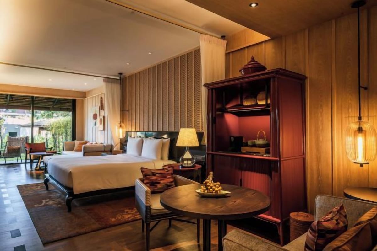 Una habitación de hotel con una cama grande, una mesa redonda con un frutero, una estantería y una lámpara.