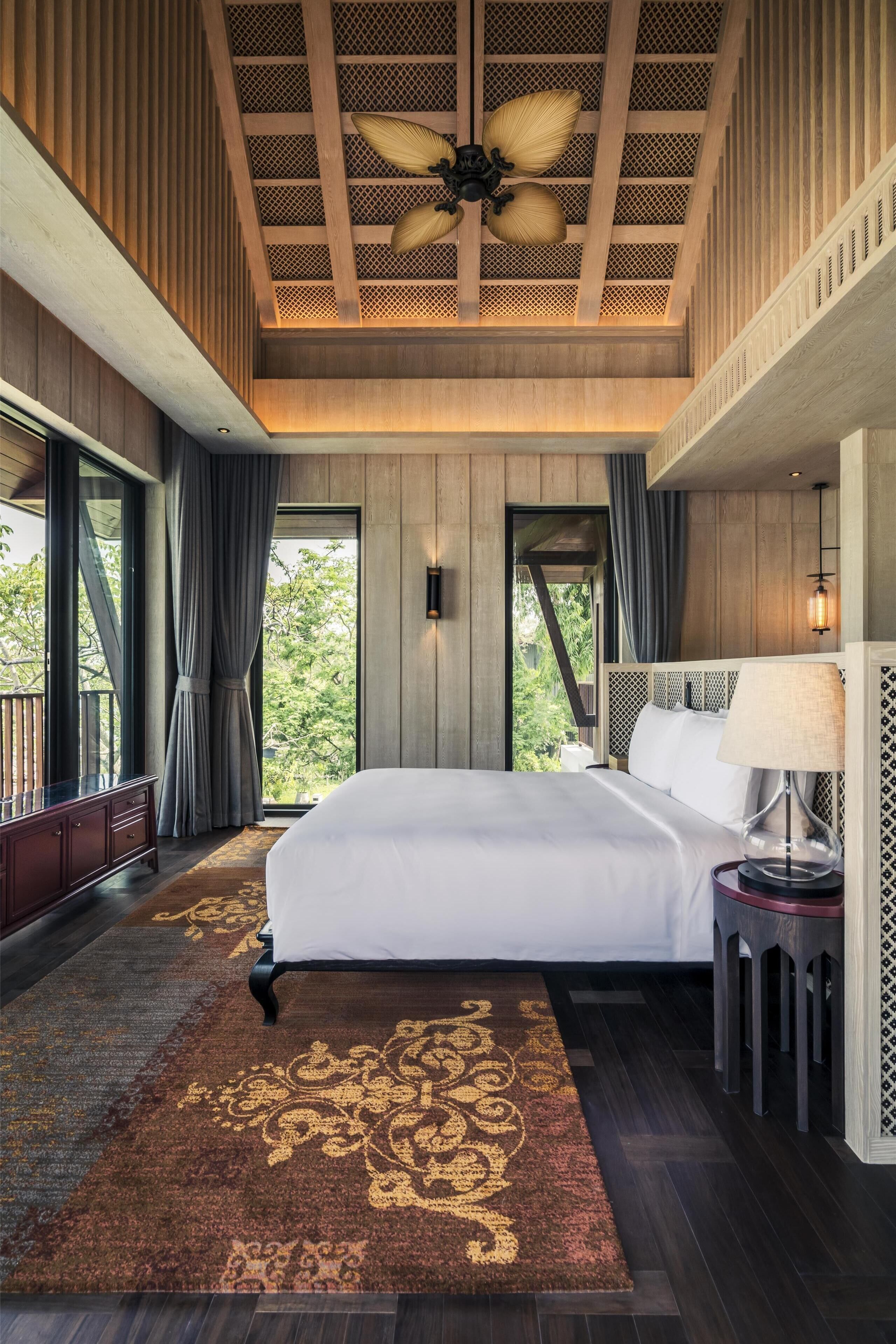 Un grand lit recouvert d’une couette blanche, un ventilateur de plafond en bois et une vue sur les arbres par la fenêtre.