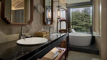 A modern bathroom with a large sink, two mirrors, a bathtub, and a window with a view of greenery.