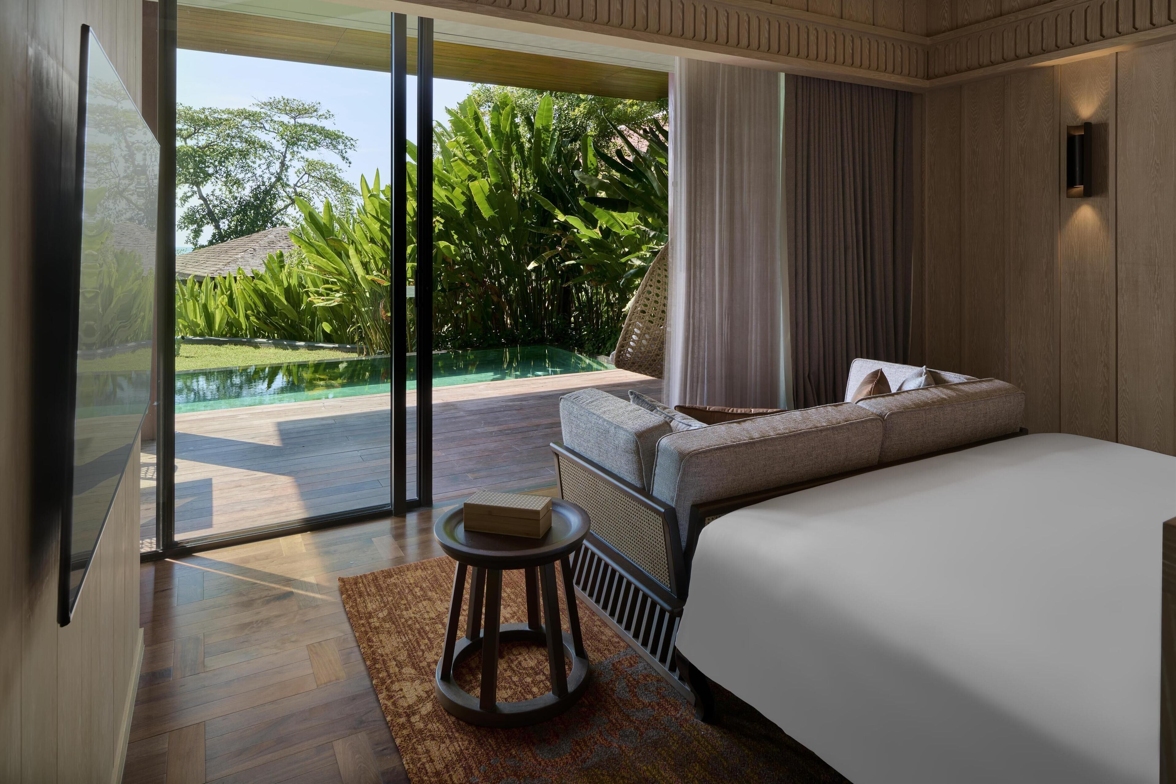 A modern bedroom with a large bed, a small stool, and a view of a swimming pool and lush greenery.