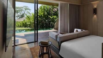 A modern bedroom with a large bed, a small stool, and a view of a swimming pool and lush greenery.