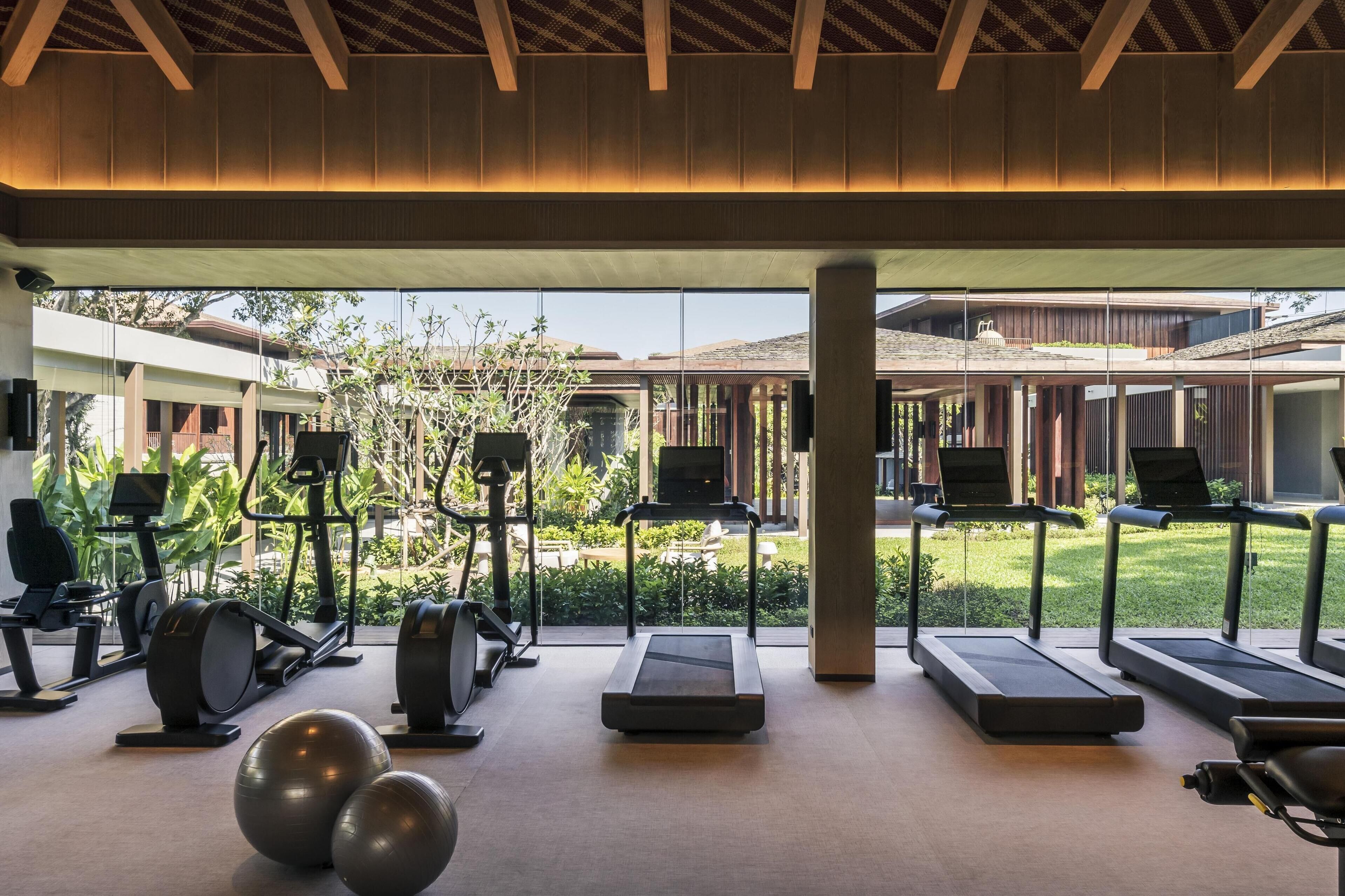 A gym with treadmills, stationary bikes, and exercise balls, with a view of a courtyard and buildings in the background.
