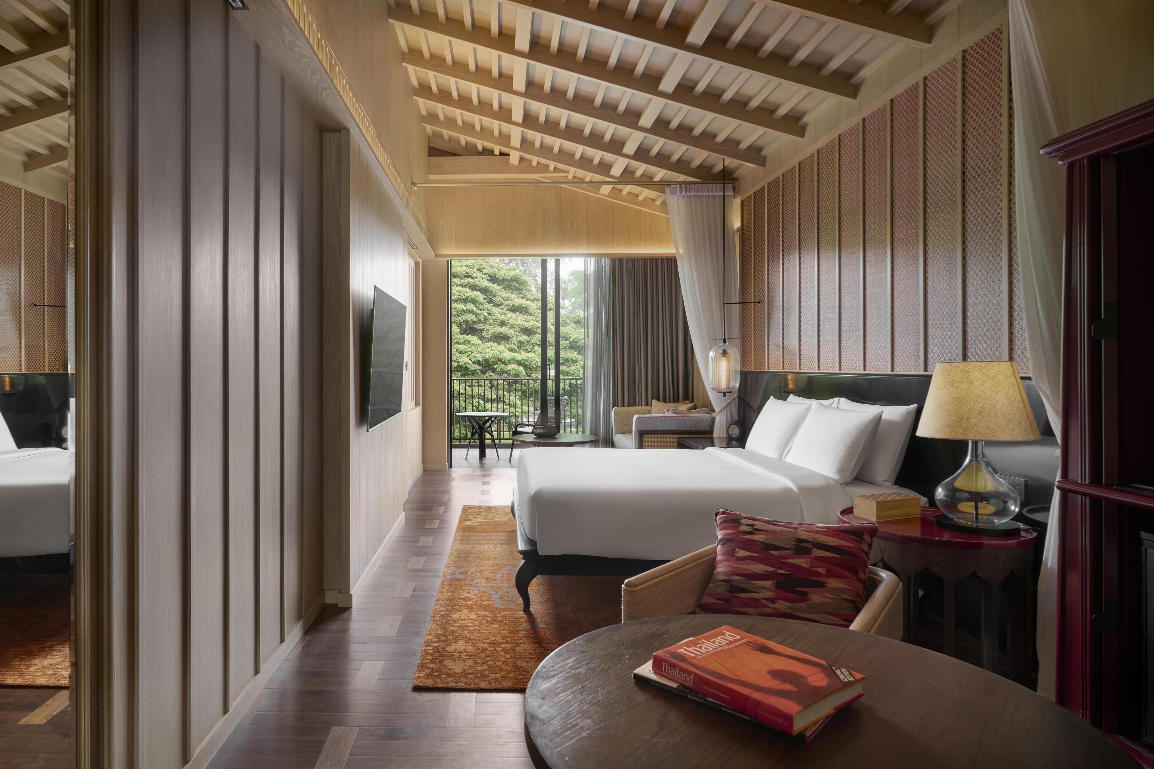 A hotel room with a bed, a desk with a lamp, a chair, a table with a book, and a view of greenery outside.