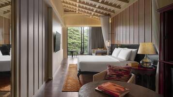 A hotel room with a bed, a desk with a lamp, a chair, a table with a book, and a view of greenery outside.