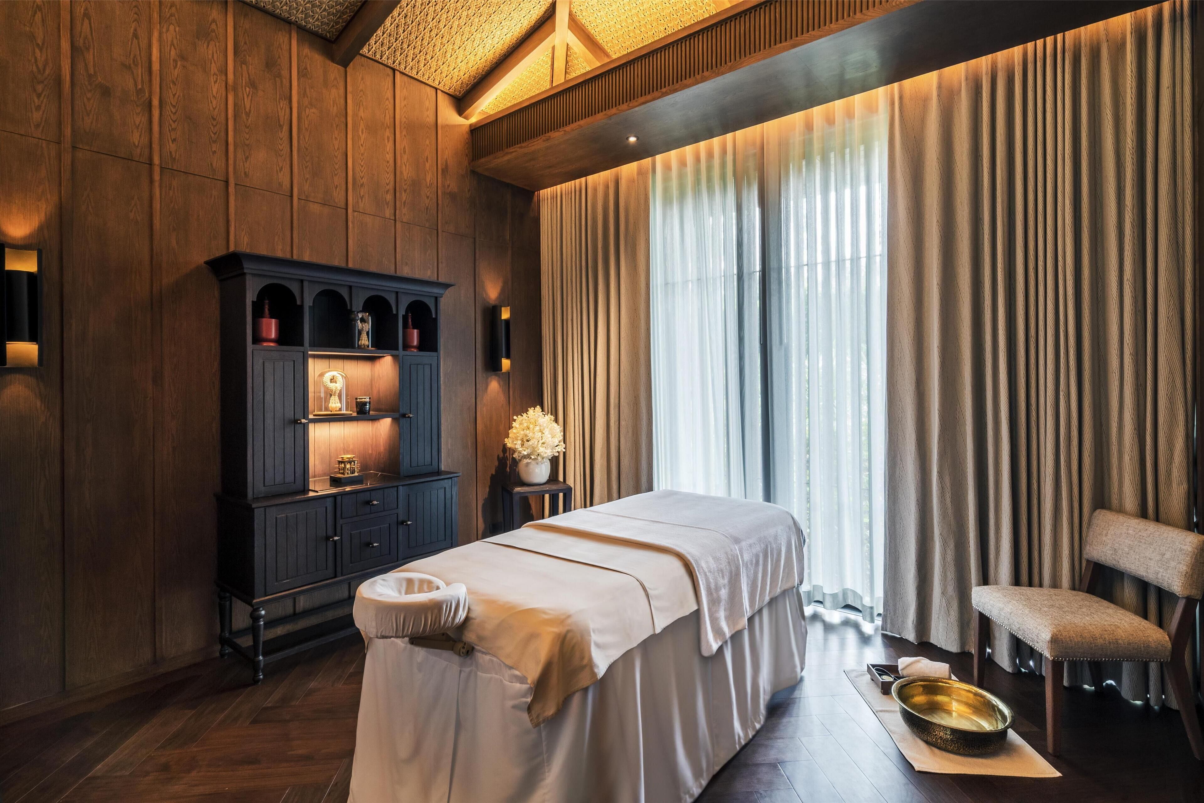 A spa room with a massage table, a cabinet with shelves, a chair, and a large window with curtains.