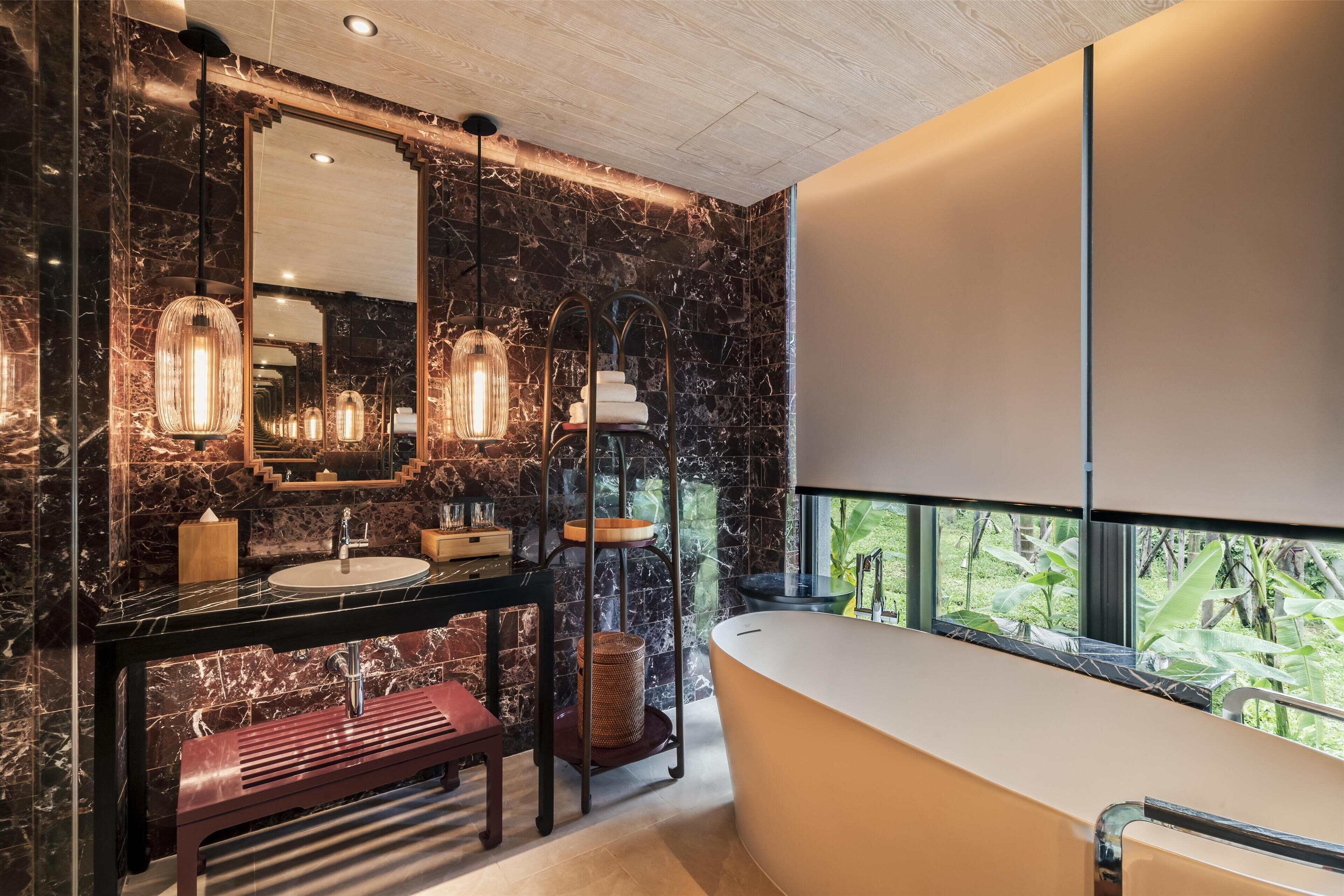 A modern bathroom with a marble wall, a large mirror, a sink, a bathtub, and a window with a view of greenery.