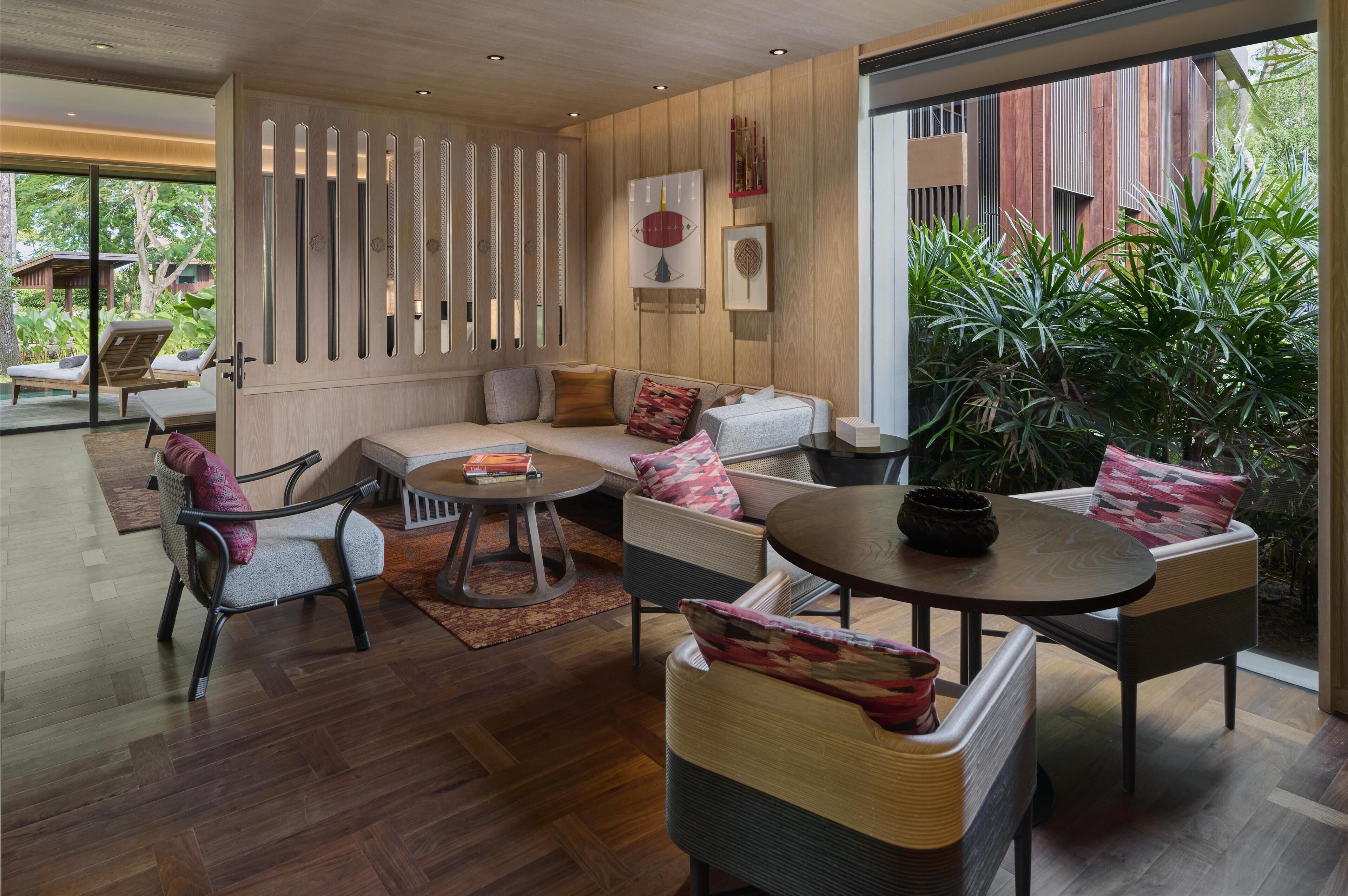 Ein modernes Wohnzimmer mit einer Couch, Sesseln und einem Couchtisch. Eine Wand aus Holzlamellen und ein großes Fenster mit Pflanzen davor.