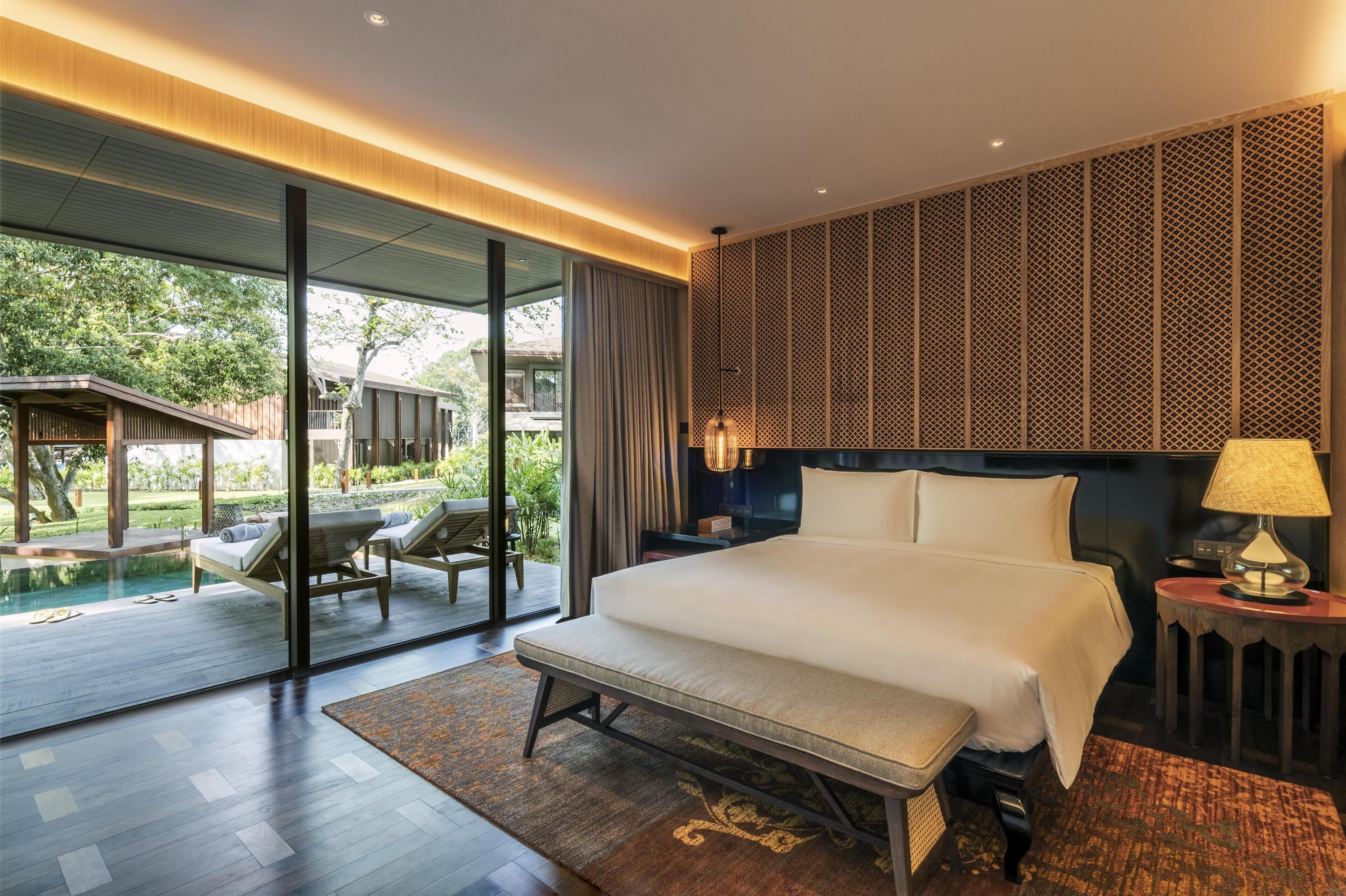 A modern hotel room with a large bed, a bench, a bedside table with a lamp, and a view of an outdoor swimming pool and garden.