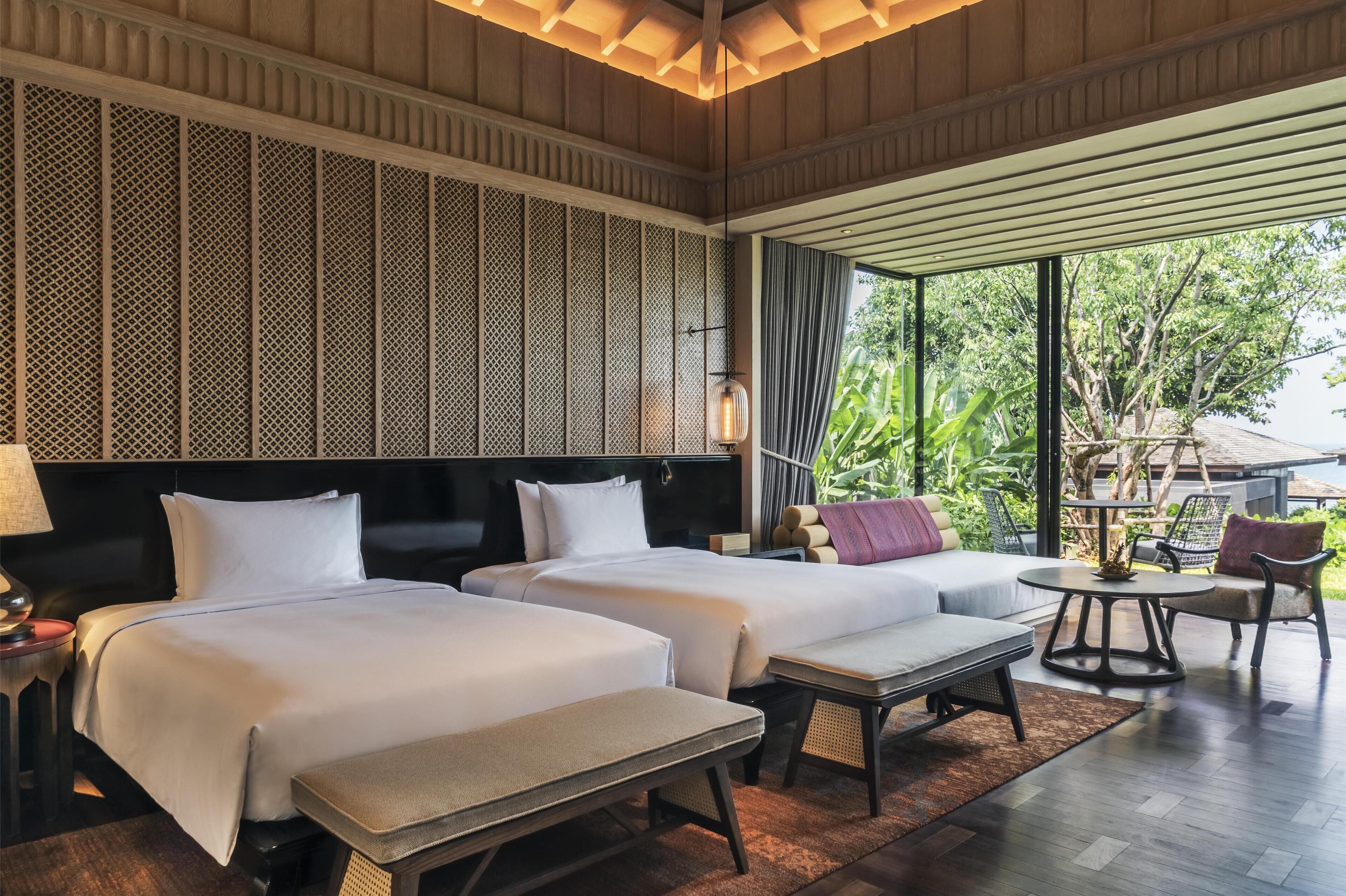 A hotel room with two beds, a sofa, a small table, and a view of greenery outside.