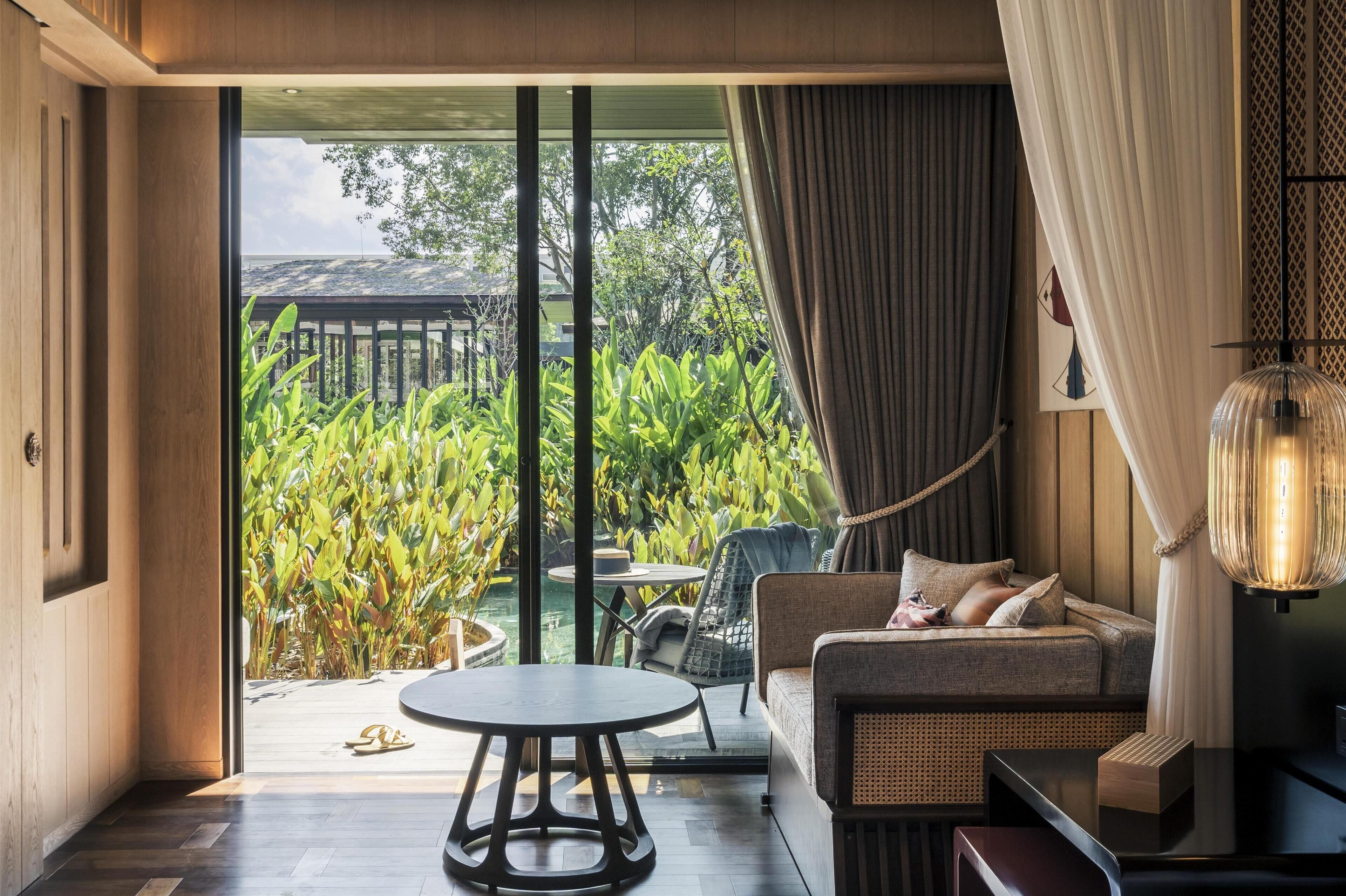 A modern living room with a sofa, a round table, and a view of a lush garden and pool.