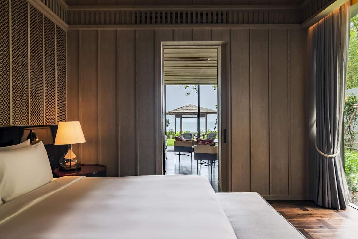 Une chambre d’hôtel avec un grand lit, une table de chevet avec une lampe et une vue sur un espace extérieur aménagé, accessible par une porte.