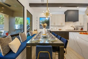 A modern dining area with a glass table, blue upholstered bench, and a large window with a view of greenery.