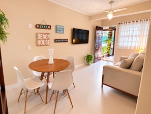 Ein Wohnzimmer mit einem runden Holztisch, weißen Stühlen, einem beigen Sofa und einem Fernseher an der Wand.