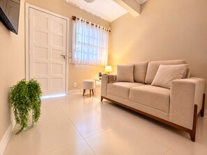 A living room with a beige sofa, a small table with a lamp, a potted plant, and a door.