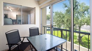 Un balcon avec une table et des chaises en verre, une vue sur les arbres et une route, et un coin cuisine visible à travers une porte vitrée.