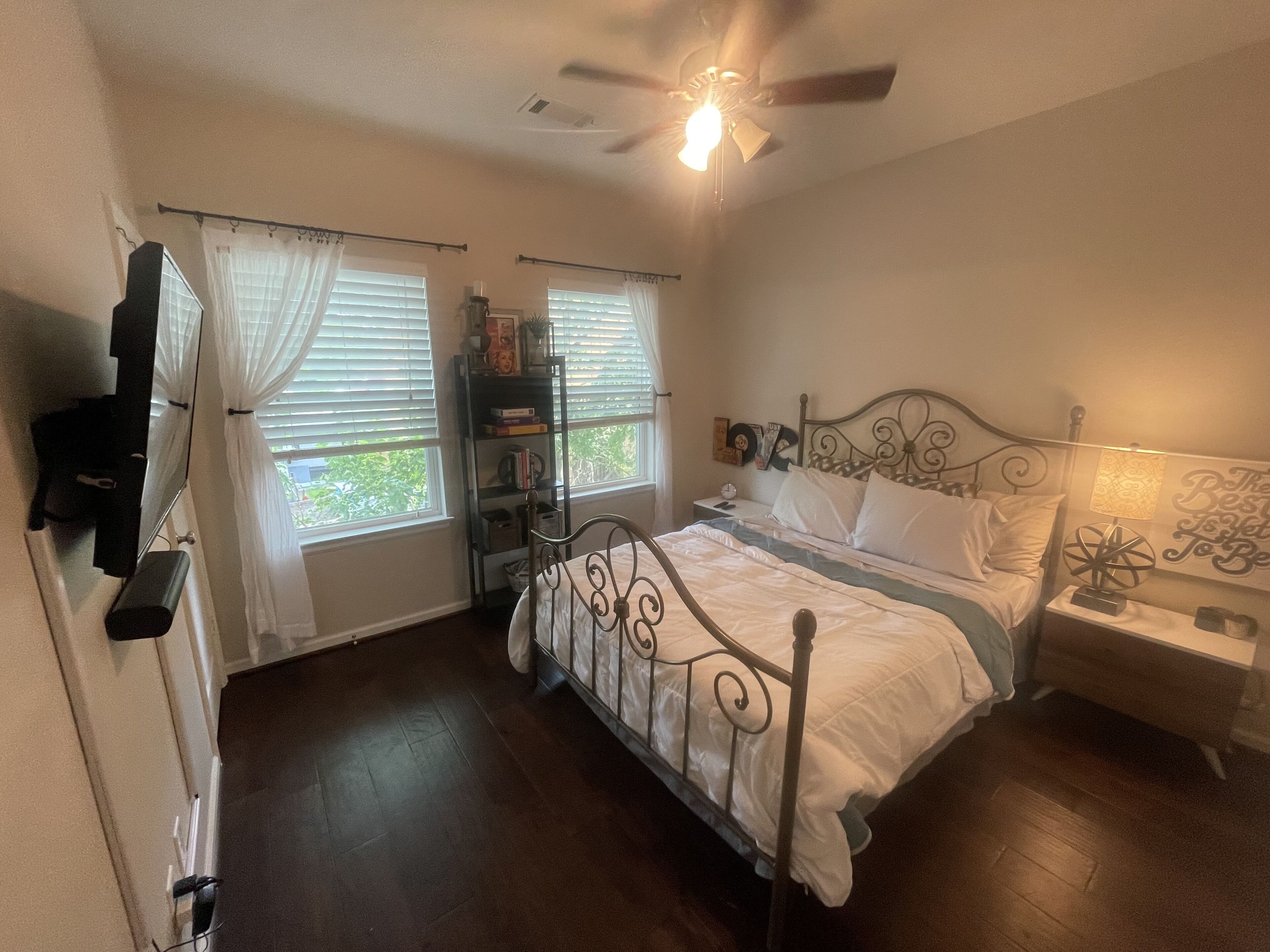 A bedroom with a bed, a ceiling fan, a nightstand, a lamp, a bookshelf, and a window with curtains.