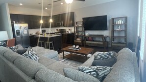 A modern living room with a grey sofa, a coffee table, and a TV. There's a kitchen area with dark cabinets and a microwave.
