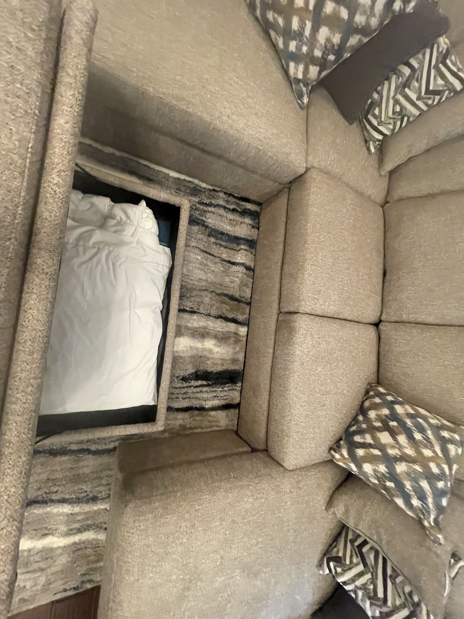 A corner of a room with a beige upholstered sofa, a patterned pillow, and a white bedspread.