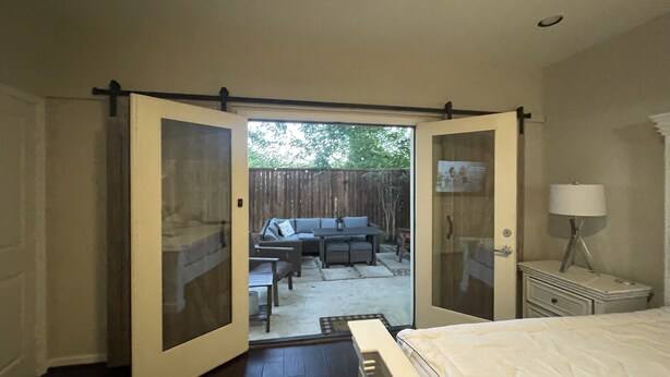 A bedroom with a bed, a nightstand, a lamp, and a sliding door leading to a patio with outdoor furniture.
