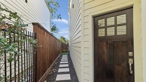 A narrow pathway leading to a dark wooden door with a window, flanked by a wooden fence and a building with white siding.