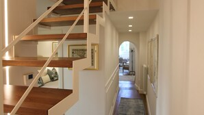 Eine Treppe mit Holzdielen und weißen Geländern führt zu einer oberen Etage. Ein blauer Teppich bedeckt den Boden, und an den Wänden hängen gerahmte Bilder.