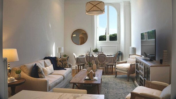 Ein modernes Wohnzimmer mit einer Couch, Couchtisch, Esstisch und Stühlen. Ein Fernseher steht auf einem Schrank, ein Spiegel ist an der Wand und ein großes Fenster sorgt für natürliches Licht.