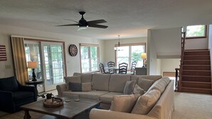 Ein Wohnzimmer mit einer Couch, einem Couchtisch und einem Deckenventilator. Eine Treppe befindet sich rechts und im Hintergrund ein Essbereich.
