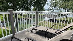 Eine Terrasse mit zwei Liegestühlen und einem weißen Geländer, das auf ein Gewässer und eine Rasenfläche blickt.