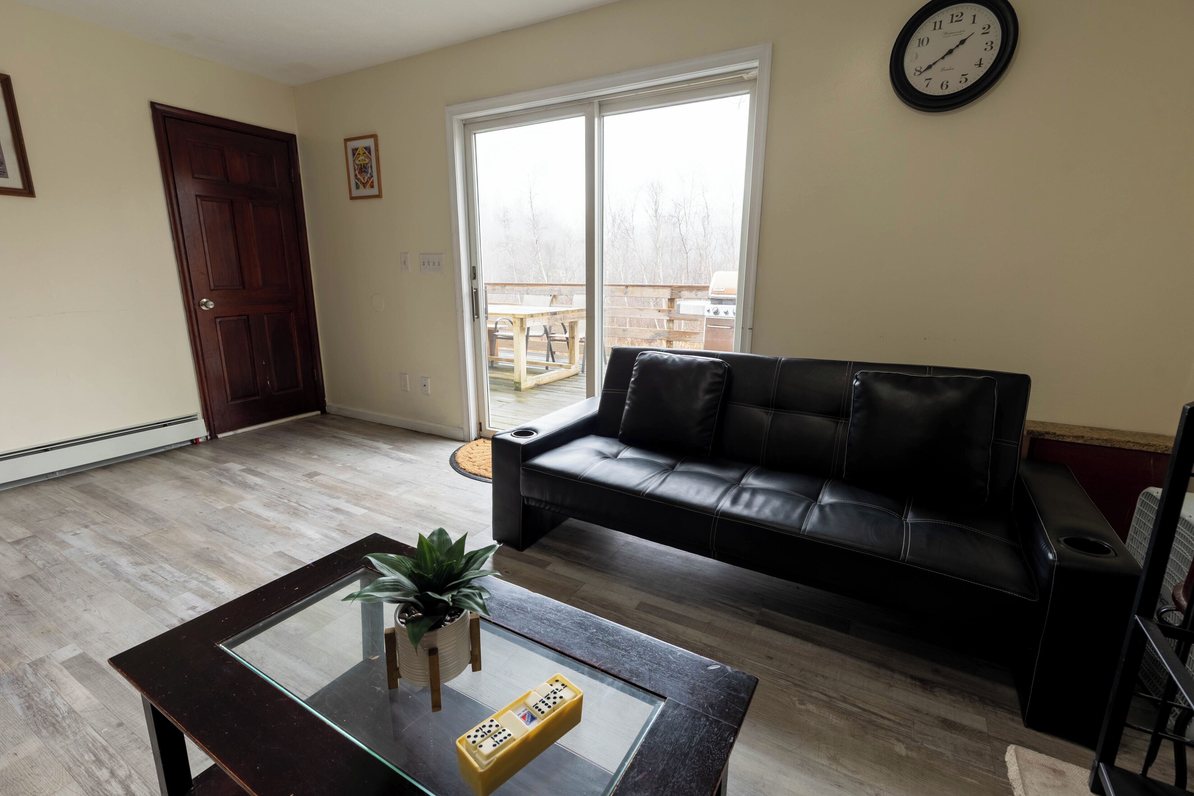 A living room with a glass-top coffee table, a black leather sofa, a wooden floor, a sliding glass door leading to an outdoor patio, and a wall clock.