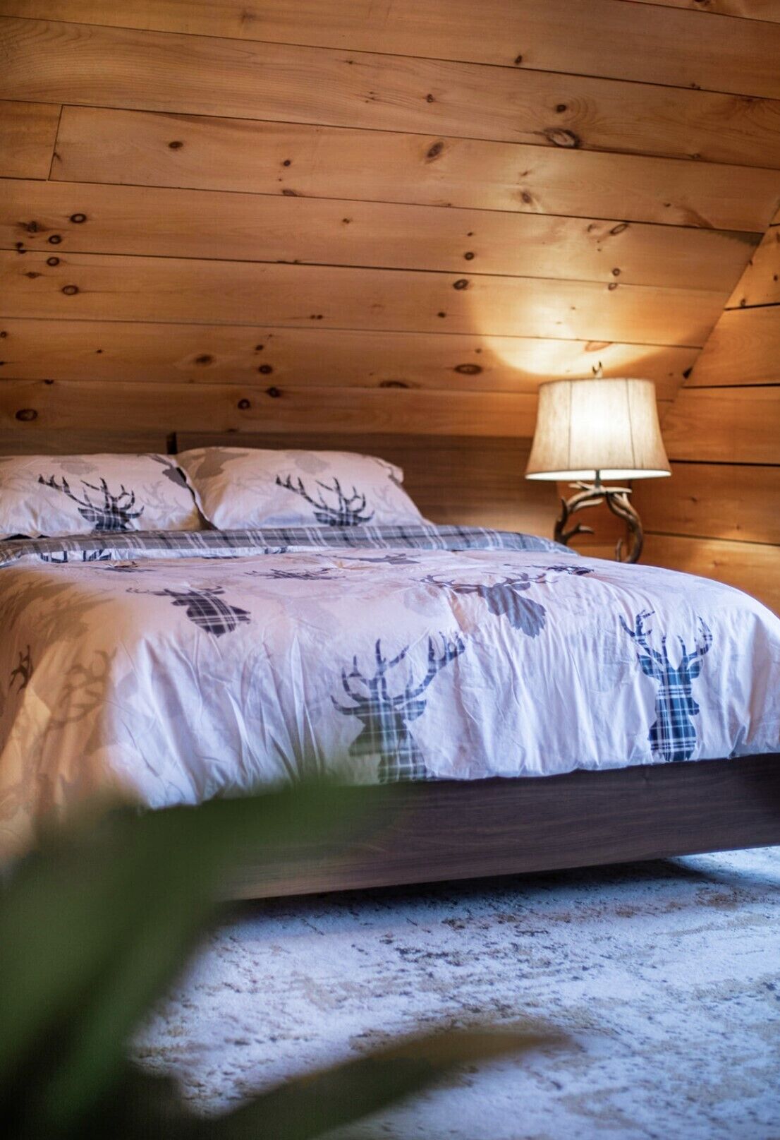 Une chambre confortable dans un chalet en bois, avec un lit recouvert d’une courtepointe à motifs de cerfs, une table de chevet avec une lampe et un plafond en bois rustique.