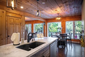 Interior de uma cabana de madeira, com cozinha, área de jantar e grandes janelas com vista para uma floresta.