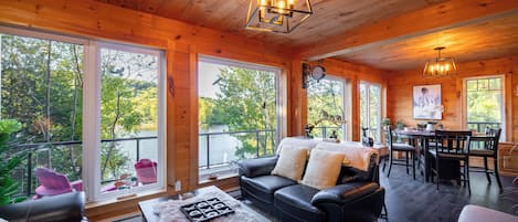 A cozy cabin interior with a dining area, a fireplace, and large windows overlooking a forest.