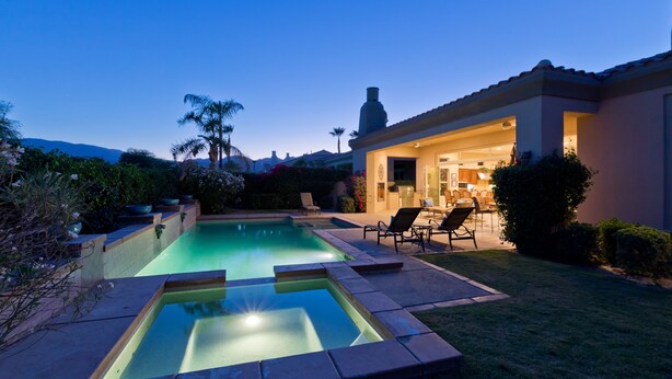 A swimming pool with a hot tub, surrounded by a patio with chairs and a covered area with a table and chairs.