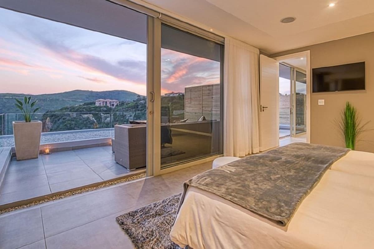 A bedroom with a large bed, a sliding glass door leading to a balcony, and a flat-screen TV mounted on the wall.