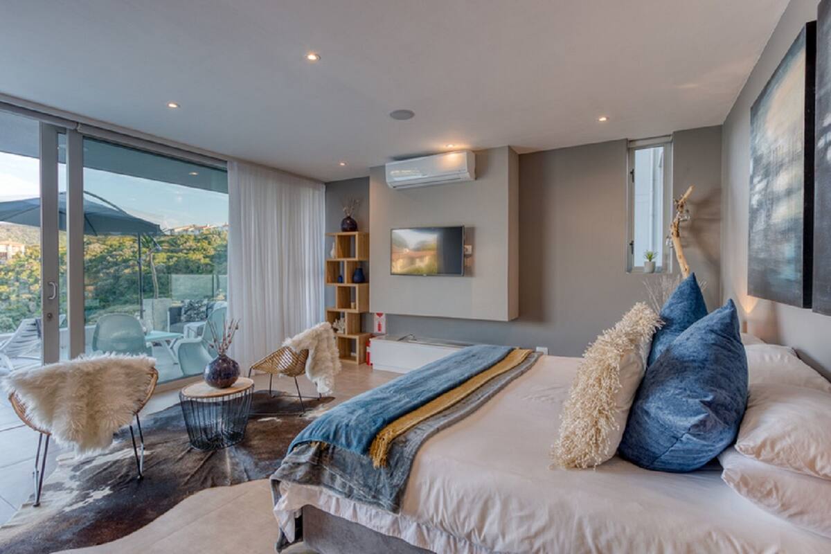 A modern bedroom with a large bed, a view of the outdoors, a built-in shelf, and a mounted artwork.