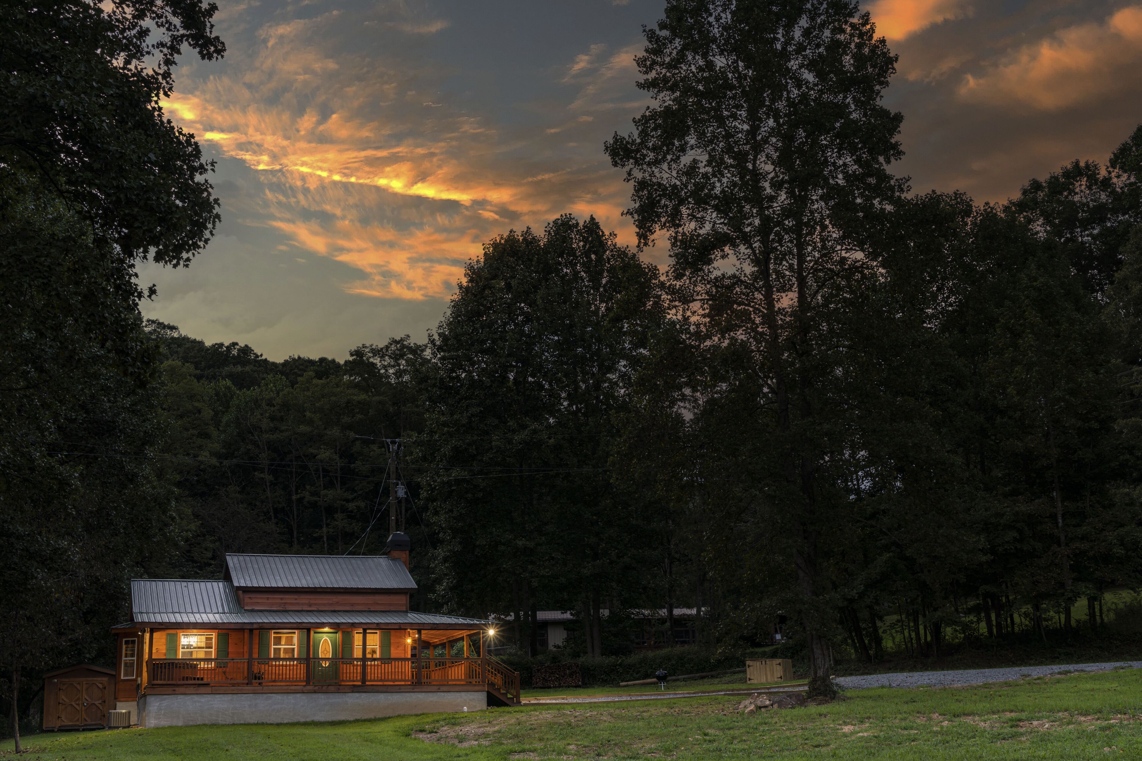 Une cabane dotée d’un porche, entourée d’une forêt dense, avec un coucher de soleil en arrière-plan.
