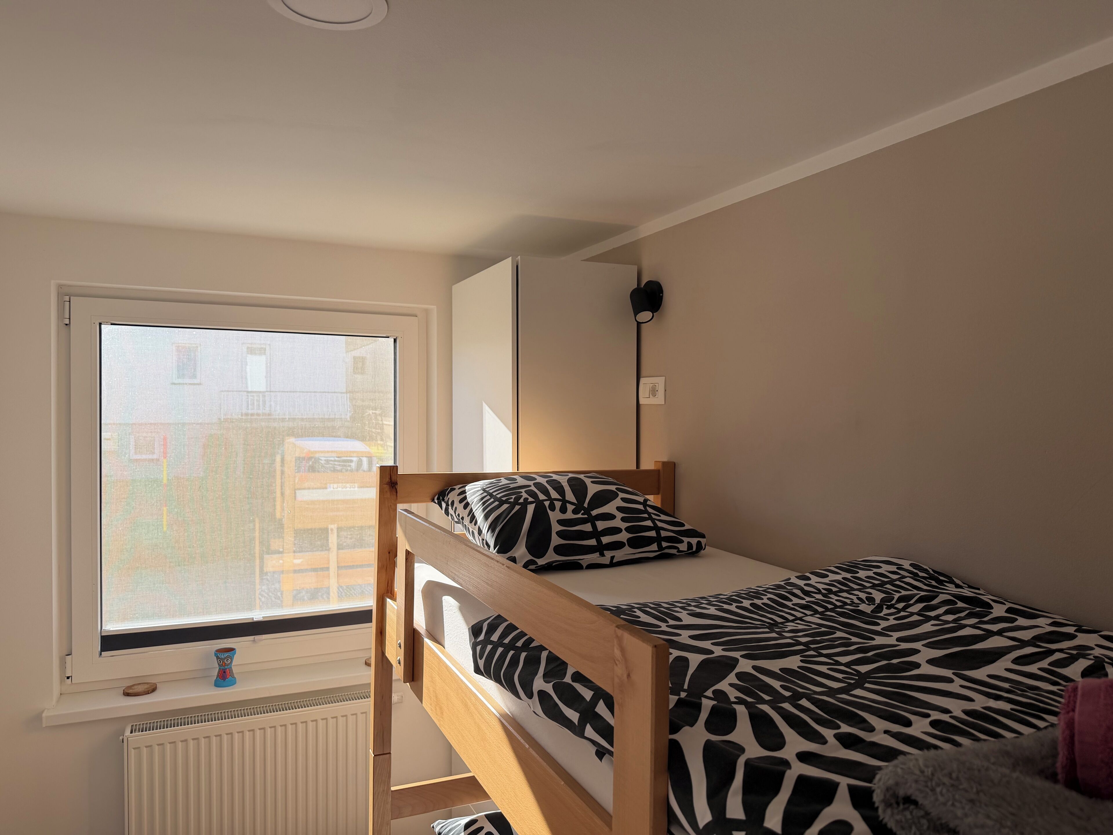 A bedroom with a bunk bed, a window with a view of buildings, a radiator, and a small table with a decorative item.