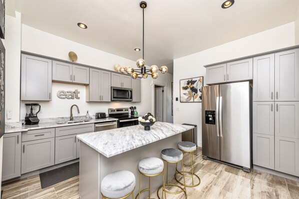 A modern kitchen with grey cabinets, a central island with stools, stainless steel appliances, and a chandelier.