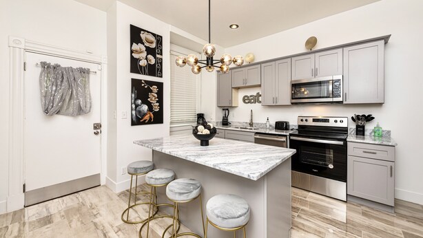 A modern kitchen with grey cabinets, a central island with stools, a chandelier, and a microwave.