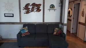Un sofà seccional gris fosc amb dos coixins decoratius, un timó de vaixell i un mural de peixos a la paret.